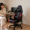木目の仕事部屋で、黒と赤のレーシング調ゲーミングチェアを見つめながら検討する女性