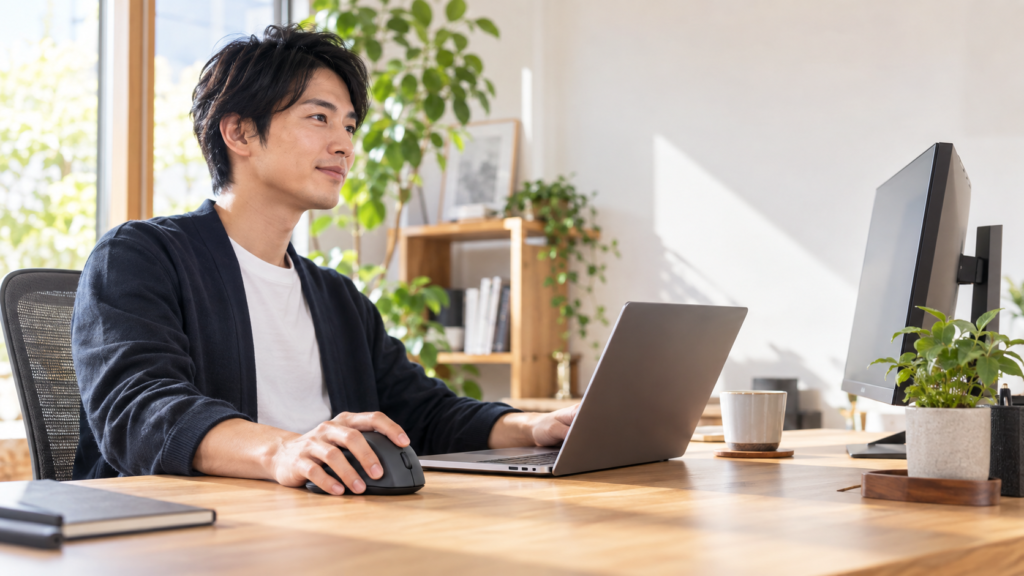 明るいデスクでノートPCと外部モニターを使いながら、ワイヤレスマウスを操作する男性