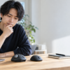 ノートPCの横で、木製デスク上の親指操作型トラックボールと通常マウスを並べて見比べる男性