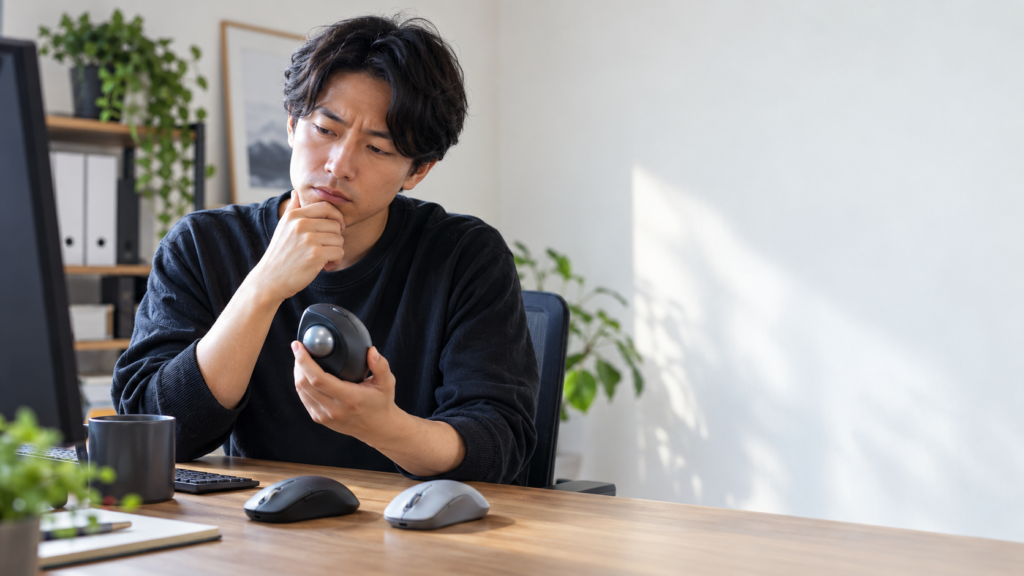 複数の通常マウスが置かれたデスクで、親指操作型トラックボールを手に取り形状を確認する男性