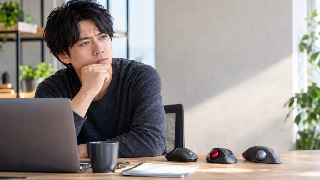 ノートPC脇に通常マウスと赤・銀のボールを備えたトラックボールを並べ、選択肢を見比べる男性